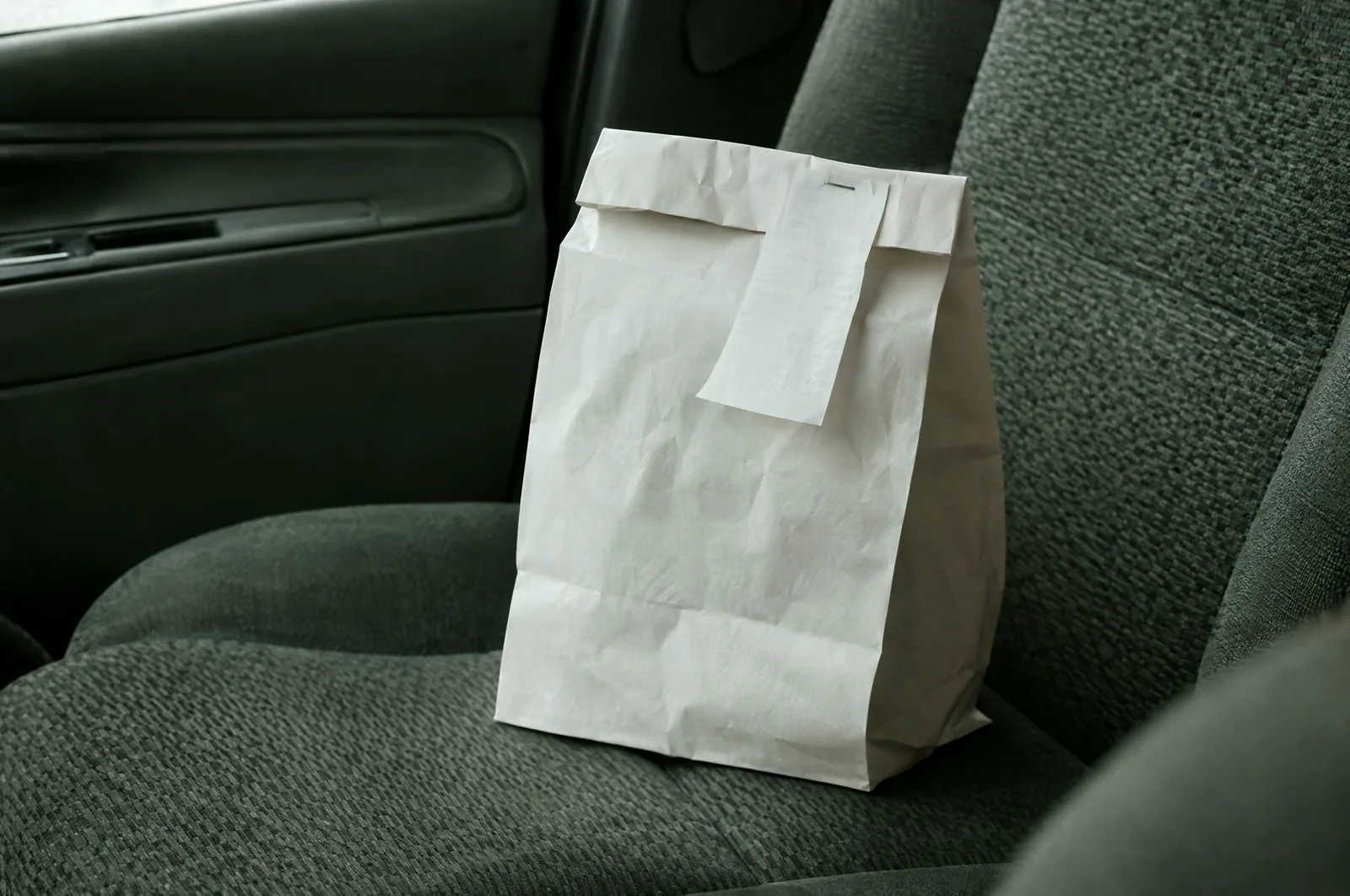 A plain white paper bag with its top folded over and a small receipt stapled to the fold, resting on a car's fabric seat.